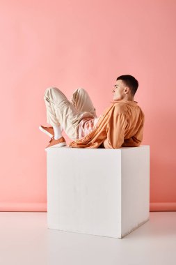 Pembe arka planda beyaz küpün üzerinde duran şeftali rengi kıyafetli bir adamın yan görüntüsü.