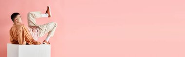 Pembe arka planda beyaz küpün üzerinde yatan şeftali rengi kıyafetli bir adamın yan görüntüsü.