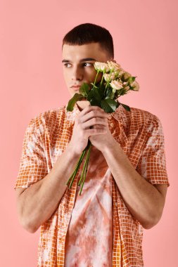 Handsome man in layered peach color outfit holding roses near face on pink backdrop clipart