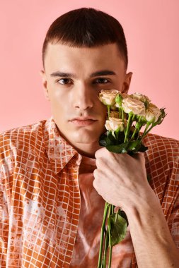 Fashionable man in layered peach color outfit holding roses near face on pink backdrop clipart