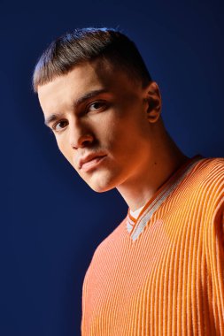 Turuncu gömlekli ve beyaz şortlu, koyu mavi arka planda poz veren yakışıklı bir adamın moda portresi.