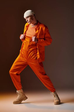 Turuncu tulumlu ve ceketli, kahverengi arka planda duran bej bereli şık bir adamın moda fotoğrafı.