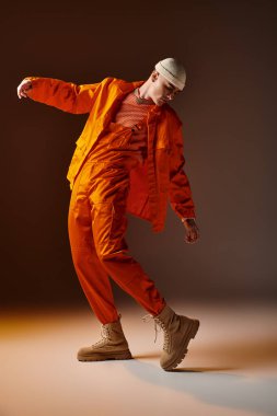 Full length image of man in orange jumpsuit and jacket, beige beanie posing on brown background clipart