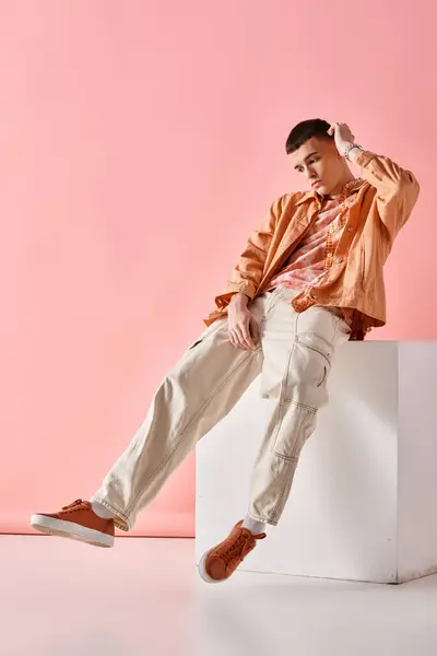 Stylish man in beige outfit touching his hair and sitting on white cube on pink background