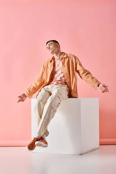 Stylish man in beige outfit holding arms open and sitting on white cube on pink background