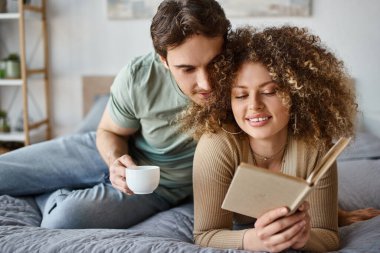 Morning cuddles and book time for curly young woman as brunette man lovingly embraces her clipart