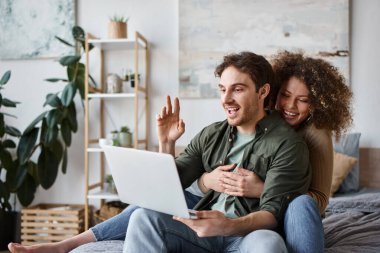 In their cozy bedroom, curly young woman and brunette man talking online on their laptop clipart