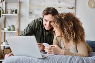 Kıvırcık genç kadın ve esmer adam yan yana çalışıyorlar laptoplarında yatak odasında gülümseyerek.