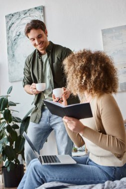 Portrait of brunette man with coffee cups looking with smile at his girlfriend working on laptop clipart