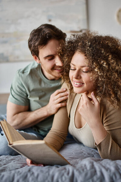 curly young woman finds comfort in her book as brunette man lovingly hugs her from behind in bed