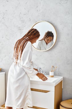 Afro örgülü Afrikalı bir kadın modern banyosunda duruyor, güzellik ve hijyen ritüelleri ile meşgul..