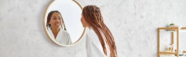 An African American woman with afro braids gazes at her reflection in a mirror in a modern bathroom, contemplating beauty and hygiene. clipart