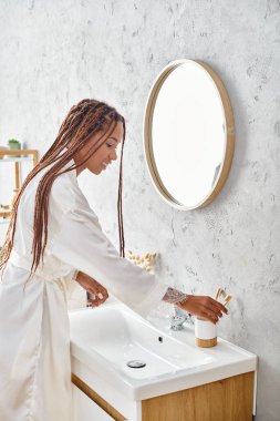 An African American woman with afro braids washes her hands in a modern bathroom, practicing personal hygiene and self-care. clipart