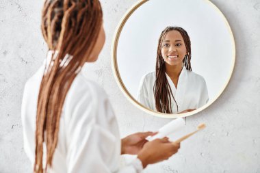 Afro örgülü bir Afro-Amerikalı kadın modern bir banyoda aynanın önünde duruyor, banyo cübbesi giyiyor..