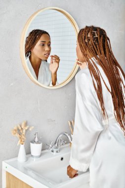 Afro örgülü Afrikalı bir kadın banyo aynasında dişlerini fırçalarken banyo cübbesi giyiyor..