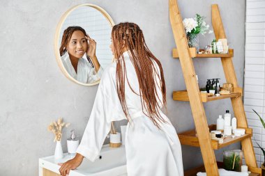 An African American woman with afro braids stands in a modern bathroom, brushing her hair in front of a mirror. clipart