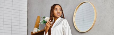 Rasta saçlı Afrikalı Amerikalı bir kadın modern bir banyoda aynanın önünde duruyor ve görünüşüne hayran..