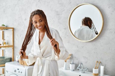 Banyo cüppeli Afro-Amerikan kadın modern banyo lavabosunun önünde duruyor, yansımasına hayran..