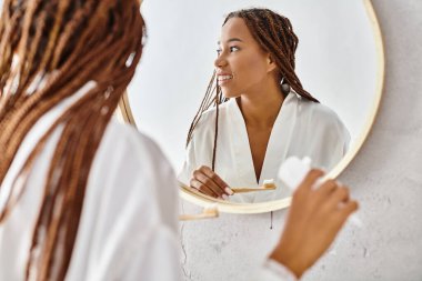 Afro örgülü bir bornoz giymiş Afrikalı Amerikalı bir kadın modern bir banyoda aynanın önünde dişlerini fırçalıyor..