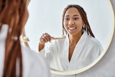 Afro örgülü, banyo cüppeli, modern banyo aynasında dişlerini fırçalayan Afrikalı bir Amerikalı kadın..