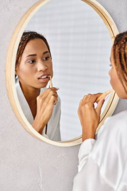 Modern banyo aynasında dişlerini fırçalayan bornozlu afro örgülü Afro-Amerikalı bir kadın..