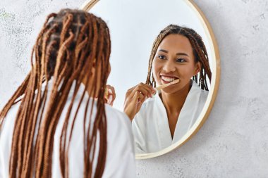Banyodaki aynanın önünde dişlerini fırçalayan banyolu afro örgülü Afro-Amerikalı kadın..