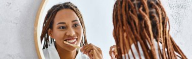 An African American woman with afro braids in a bathrobe brushes her teeth in a modern bathroom while looking at her reflection in the mirror. clipart