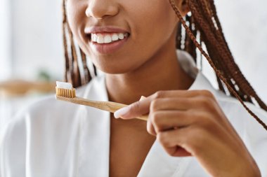 Afro örgülü bir bornoz giymiş Afrikalı Amerikalı bir kadın modern bir banyoda dişlerini fırçalıyor güzellik ve hijyen için..