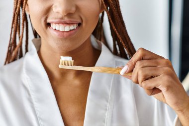 Modern bir banyoda dişlerini ahşap bir diş fırçasıyla fırçalayan bornozlu afro örgülü Afrikalı bir Amerikalı kadın..