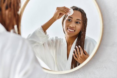 Afro örgülü Afrikalı bir kadın modern bir banyoda aynanın önünde toner kullanıyor.