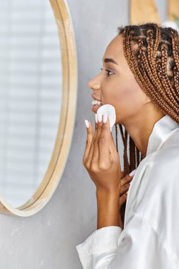 Afro örgülü, modern bir banyoda banyo bornozu giyerken toner kullanan Afro-Amerikalı bir kadın..