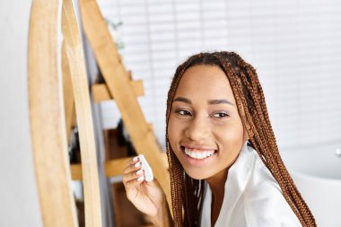 Afro örgülü Afrikalı bir kadın elinde diş fırçasıyla modern bir banyoda gülümsüyor, neşe ve hijyen saçıyor..