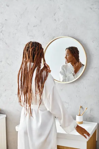 在一个现代化的浴室里 一个头发蓬乱的女人站在镜子前 穿着浴衣审视自己的美丽 — 图库照片