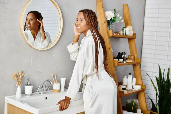一位戴着非洲式辫子的非洲裔美国妇女站在现代浴室的镜子前 — 图库照片