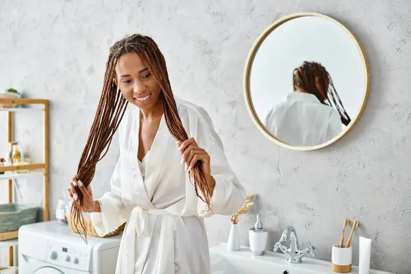 stock image African American woman with afro braids in bath robe, stands in front of modern bathroom sink, admiring her reflection.