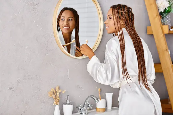 在镜子前的一个现代浴室里 一个身穿非洲式辫子浴衣的非洲裔美国女人在刷牙 — 图库照片