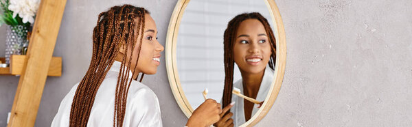 African American woman with afro braids brushes her hair in a modern bathroom wearing a bathrobe.