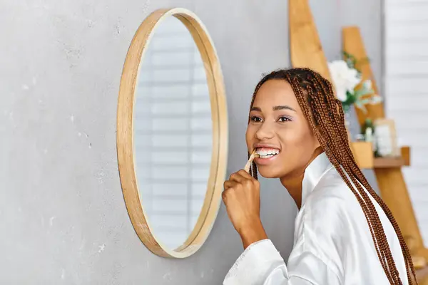 一位穿着非洲式辫子 身穿浴衣的非洲裔美国妇女在现代浴室的镜子前刷牙 — 图库照片