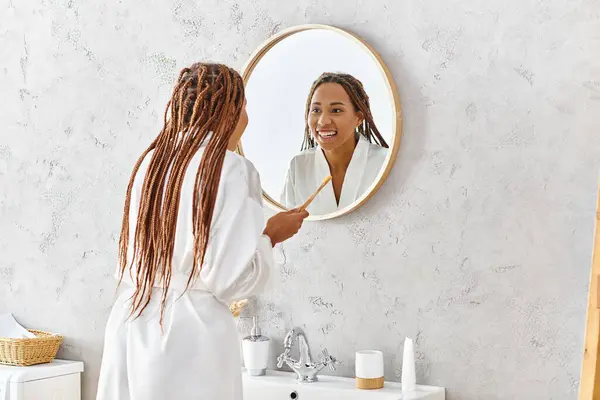 在现代浴室的镜子里 穿着浴袍刷牙的非洲裔美国女人 — 图库照片