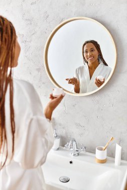 An African American woman with afro braids stands in a modern bathroom, holding beauty jar with cream clipart