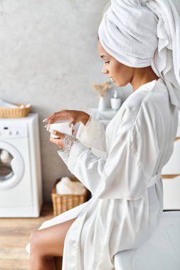 An African American woman with afro braids sits in a bathrobe and holding jar with cream in her modern bathroom. clipart