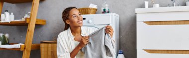 Afro örgülü Afro-Amerikalı bir kadın çamaşır makinesinin yanında bir elbise tutuyor.