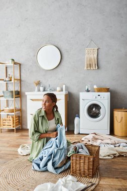 An African American woman with afro braids sitting on the floor in front of a washing machine, doing laundry in a bathroom. clipart