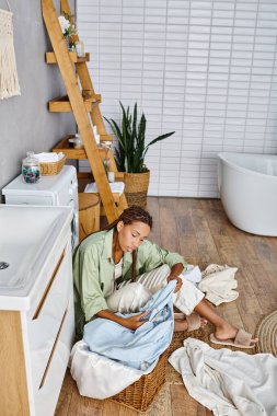 Afro örgülü Afrikalı bir kadın banyo zemininde oturuyor, ev işi yaparken etrafı çamaşırlarla çevrili..