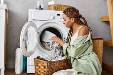 Afro örgülü Afrikalı bir kadın çamaşır makinesinin yanında oturmuş banyoda çamaşır yıkıyor..