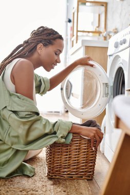 Afro örgülü Afrikalı bir kadın banyoda çamaşır makinesinin yanında oturur ve çamaşır yıkamaya odaklanır..