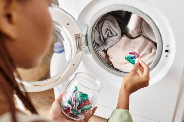 An African American woman with Afro braids gazes at gel capsule pod in a washing machine while doing laundry in a bathroom. clipart