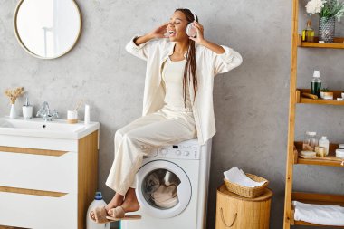 Afro örgülü Afrikalı bir kadın çamaşır makinesinin üzerinde sessizce oturur ve banyoda çamaşır yıkar..