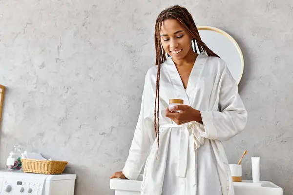 穿着浴衣的非洲裔美国女人在现代浴室里拿着装有奶油的美容罐 — 图库照片