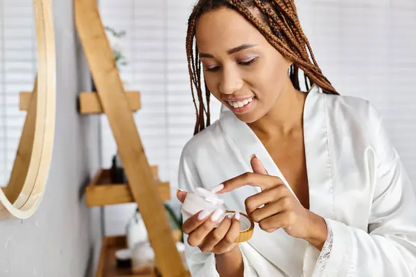 stock image An African American woman with afro braids is carefully applying cream while wearing a luxurious bathrobe in a modern bathroom.